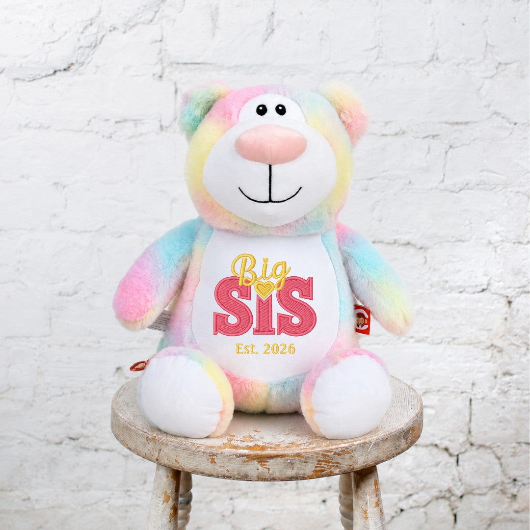 True Luv 1st pastel tie-dye plush teddy bear with Big Sis 2026 embroidery sitting on a rustic wooden stool